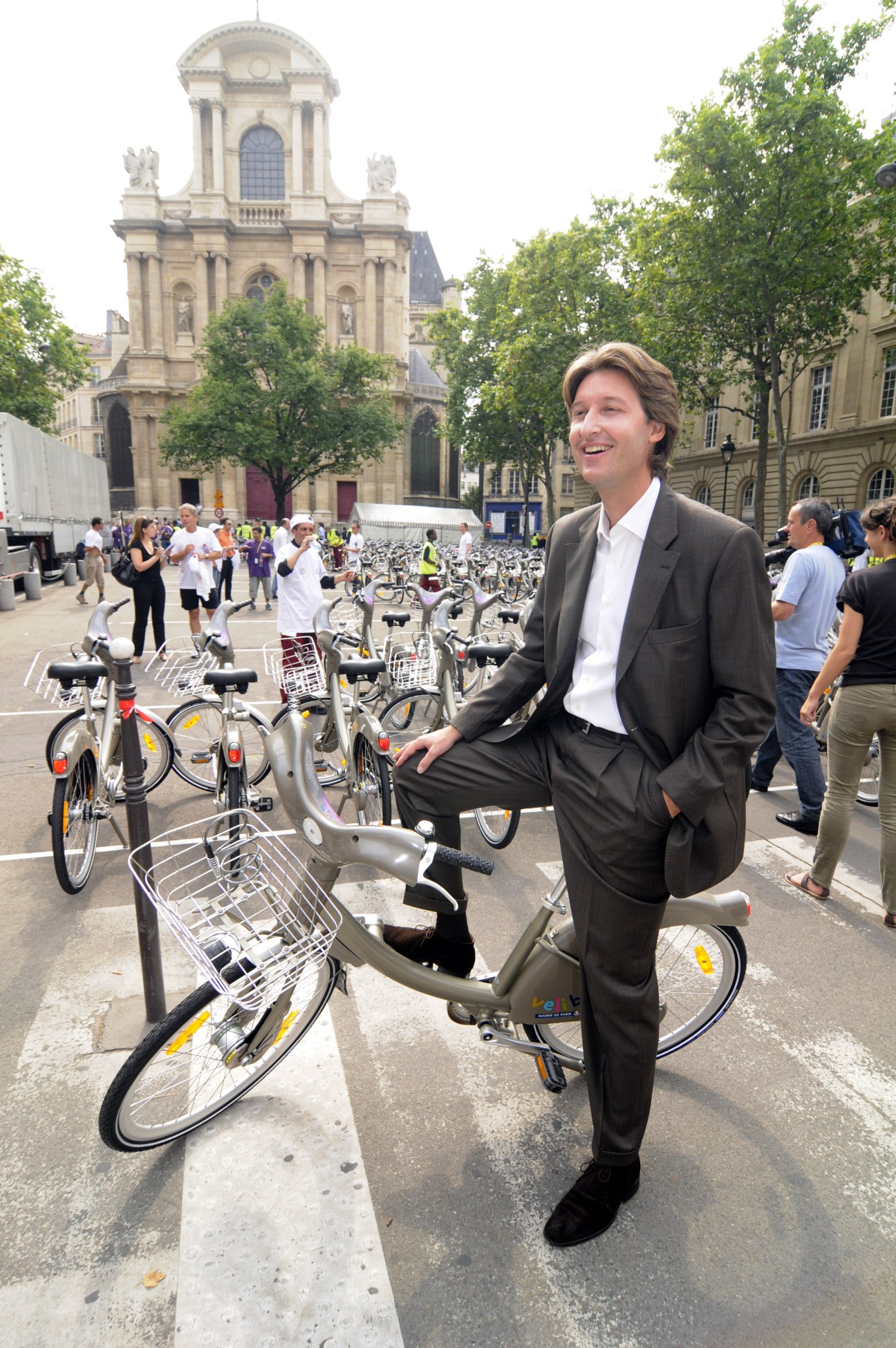 Man in business suit sitting on a Vélib' bicycle in a Paris square with a classical church building in the background.