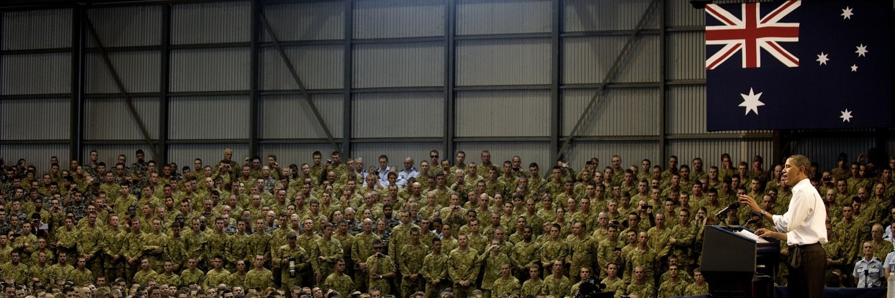 President Barack Obama addresses uniformed troops in a large hangar with Australian flag displayed.