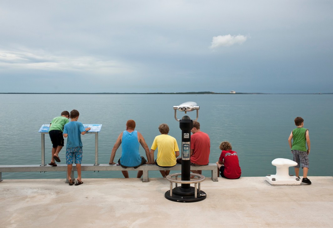 Visitors at a waterfront viewing platform with telescopes and informational displays overlooking calm bay waters.