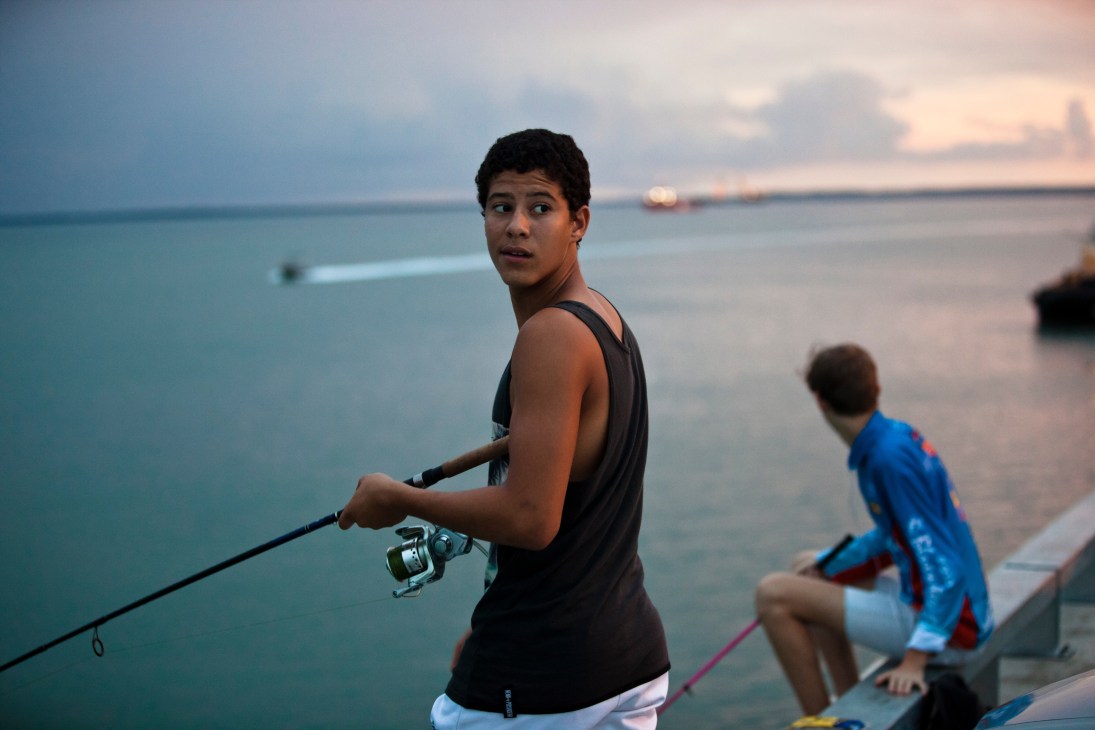 A young fisherman holding a fishing rod at Stokes Hill Wharf at dusk with calm water and boats in the background.