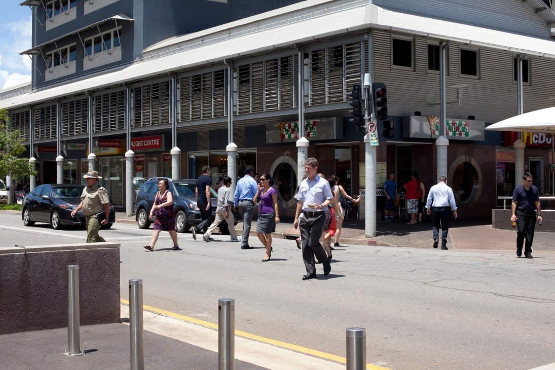 Pedestrians cross a sunny street in central Darwin with modern buildings and storefronts featuring louvered windows.