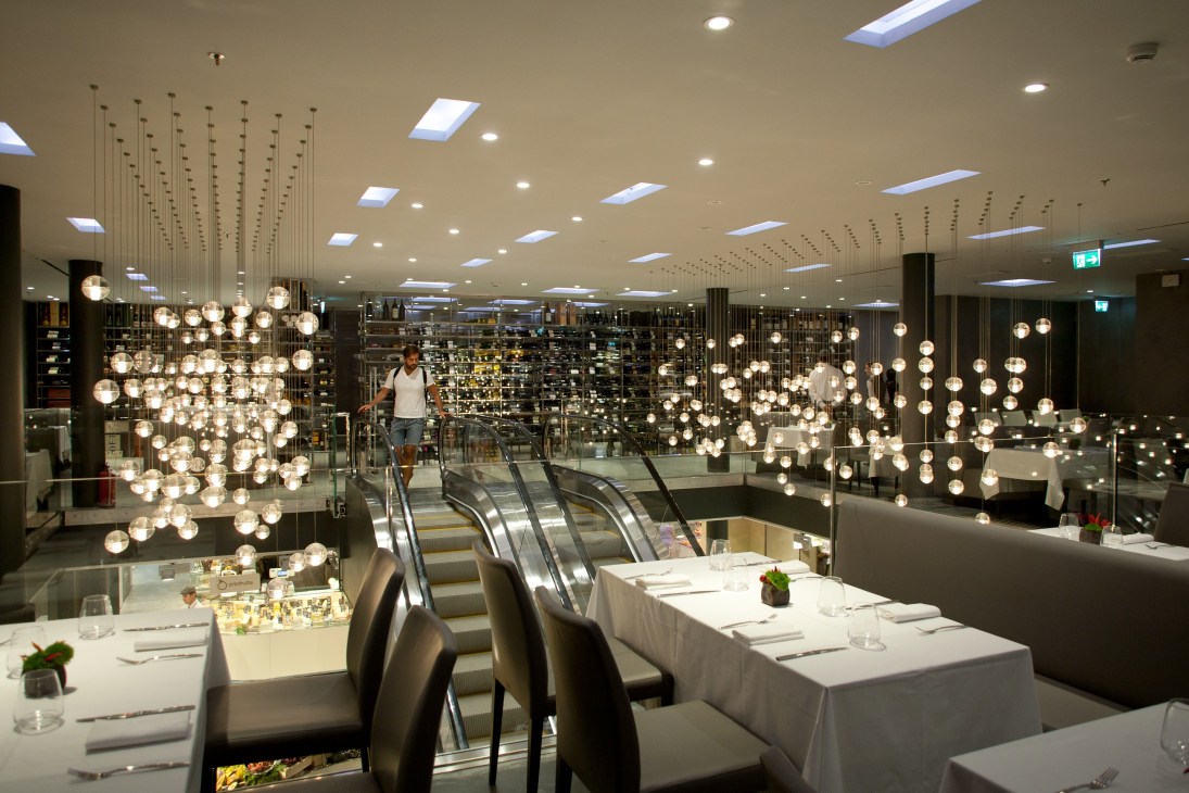 Modern restaurant interior with hanging globe pendant lights, white tablecloths, and escalators visible in background