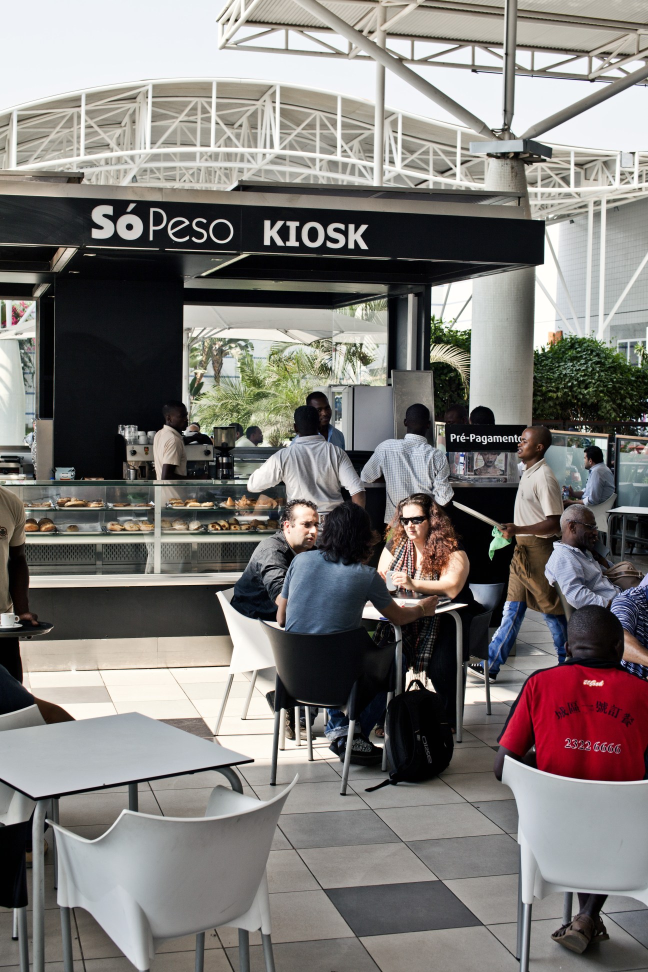 Outdoor café seating area at a shopping mall with customers dining under a metal framework structure.