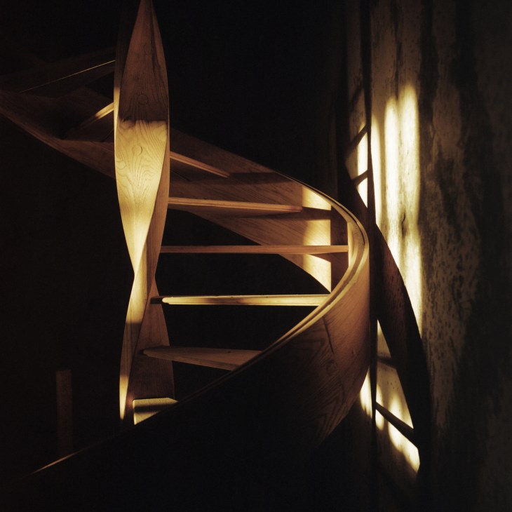 Illuminated wooden spiral staircase with curved railings against a dark background.