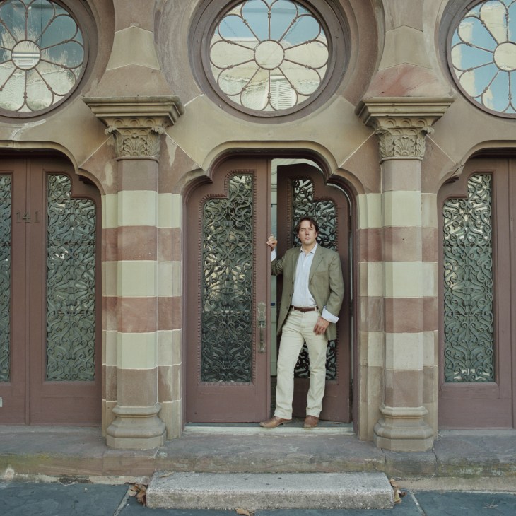 John Paul Huguley, founder of the ACBA, standing in a doorway of a historic building with ornate ironwork doors and circular windows.