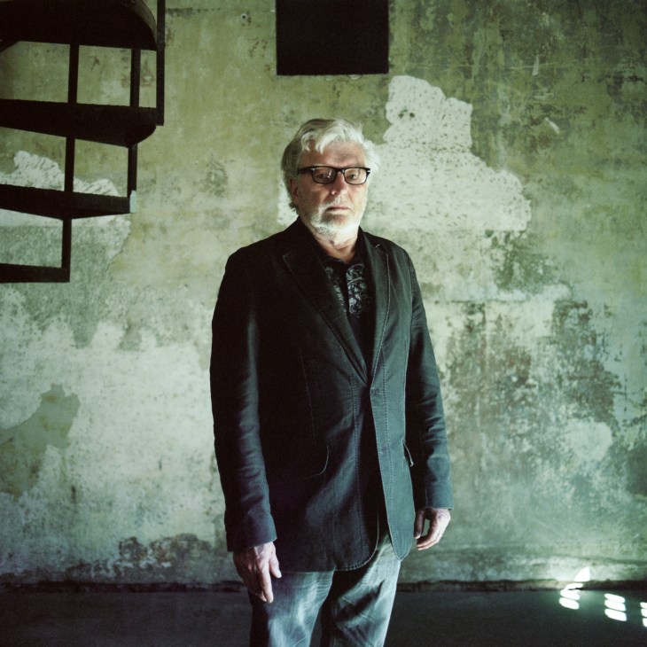 An older man with white hair and glasses stands in front of a weathered wall with a barred window.