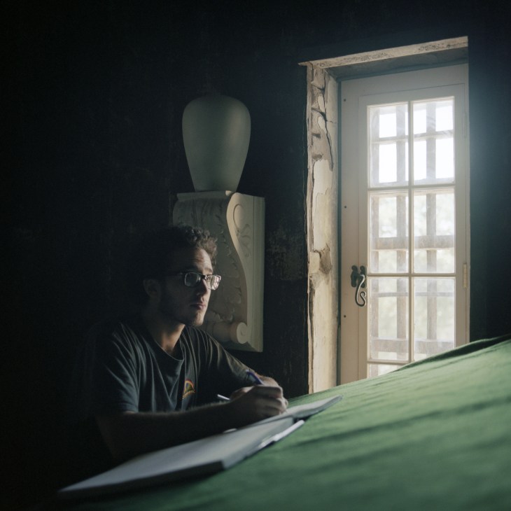 Hugh Holborn, wearing glasses, sits at a work table in a darkly lit classroom with architectural models and a tall window.