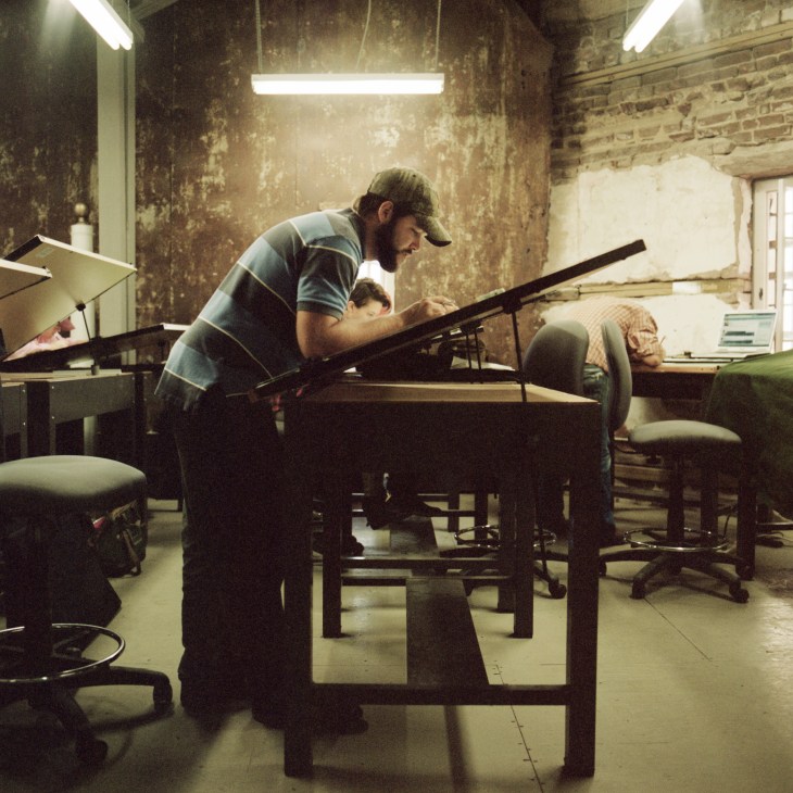 Bearded man in striped shirt working on wooden furniture construction in workshop.