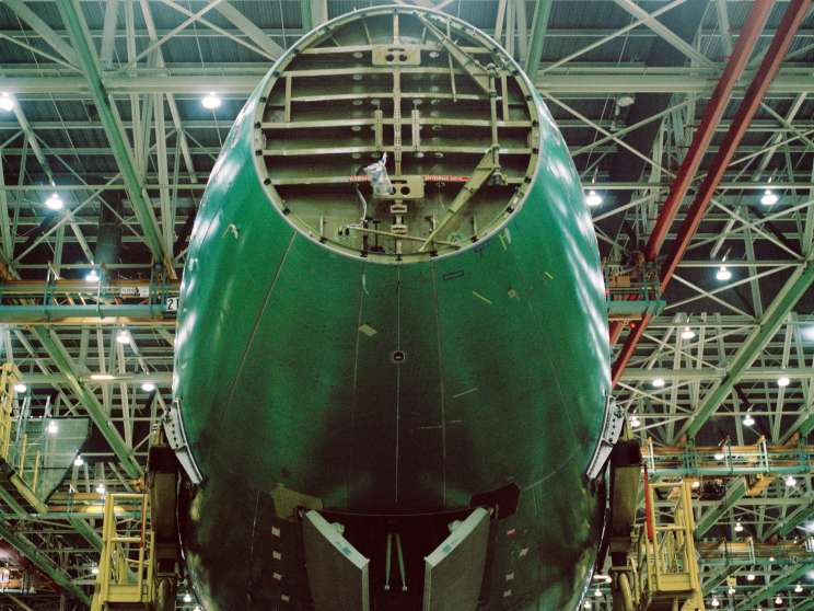 Front view of a Boeing 747-8 fuselage painted green inside a vast industrial hangar with exposed steel framework and yellow support structures.