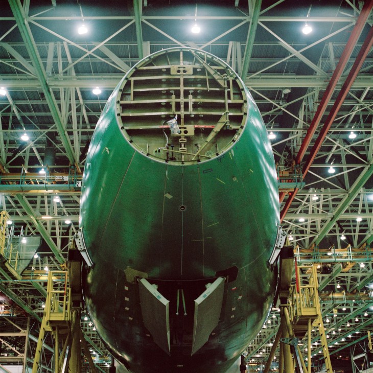 Front view of a Boeing 747-8 fuselage painted green inside a vast industrial hangar with exposed steel framework and yellow support structures.