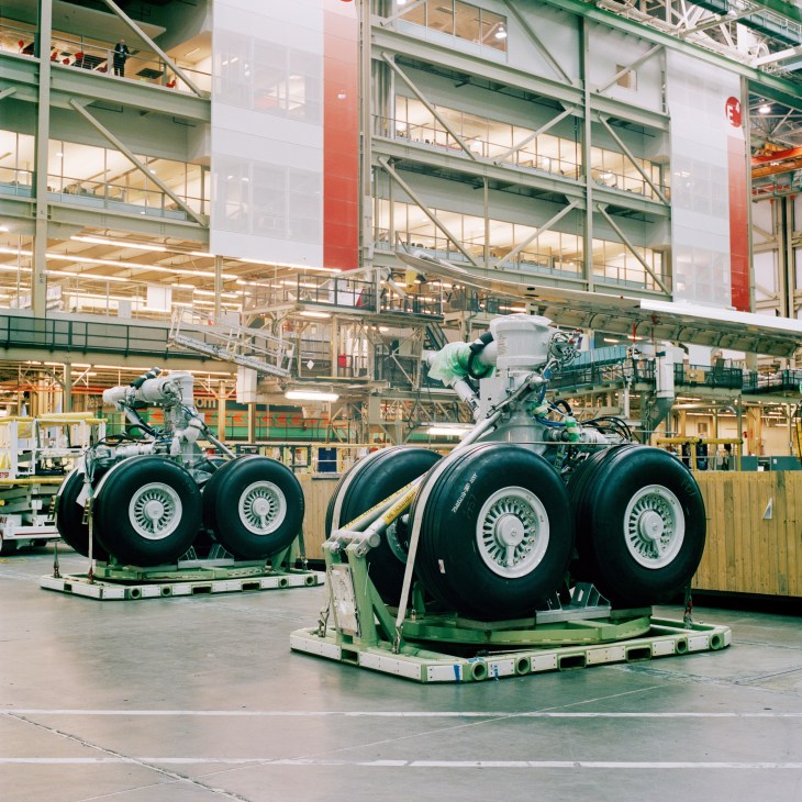 Workers install landing gear assemblies in a Boeing factory with green industrial scaffolding and overhead cranes visible.