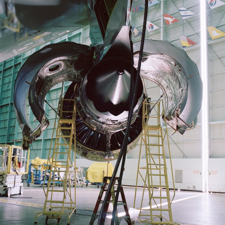 A technician works on a large aircraft engine suspended by straps in a Boeing factory, surrounded by yellow scaffolding.