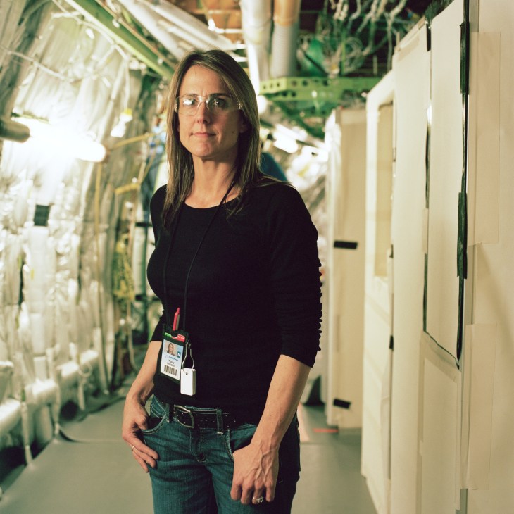 Tracy Strieby, a Boeing employee, stands in an aircraft assembly facility wearing a black top and ID badge.