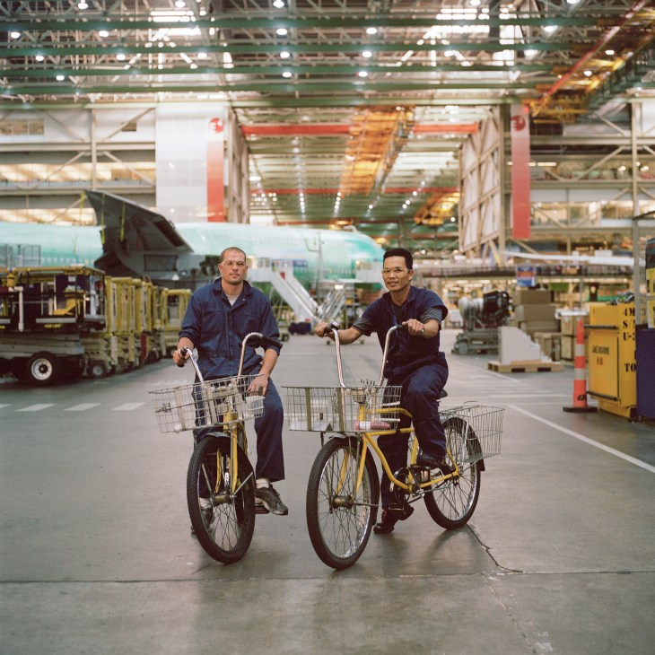 Two Boeing employees ride yellow bicycles through the vast Everett factory hangar with a 747-8 aircraft under construction in the background.