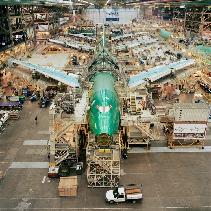 Overhead view of a green Boeing 747-8 under assembly in a massive hangar with surrounding work stations and scaffolding.