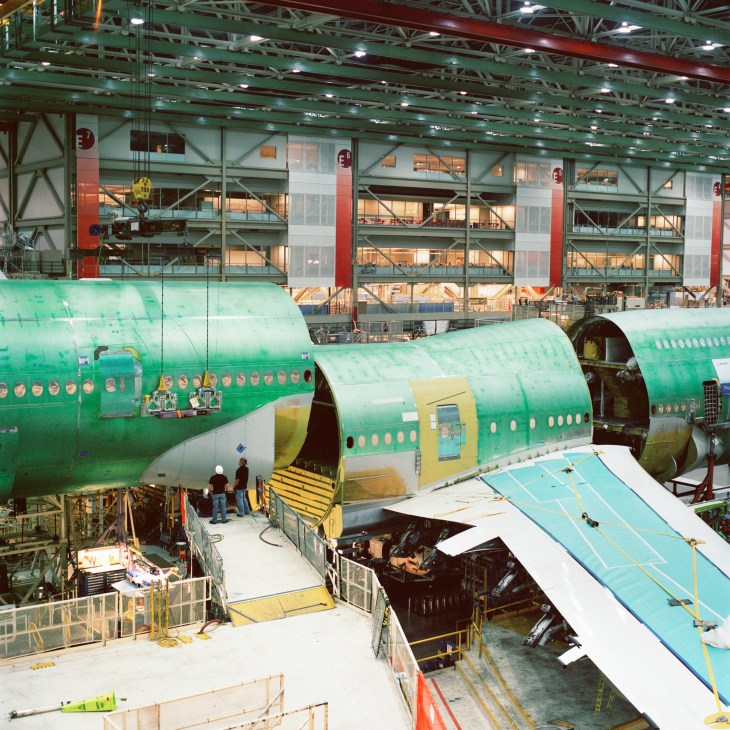 Boeing 747-8 fuselage sections in bright green being assembled in a vast factory hangar with workers nearby.