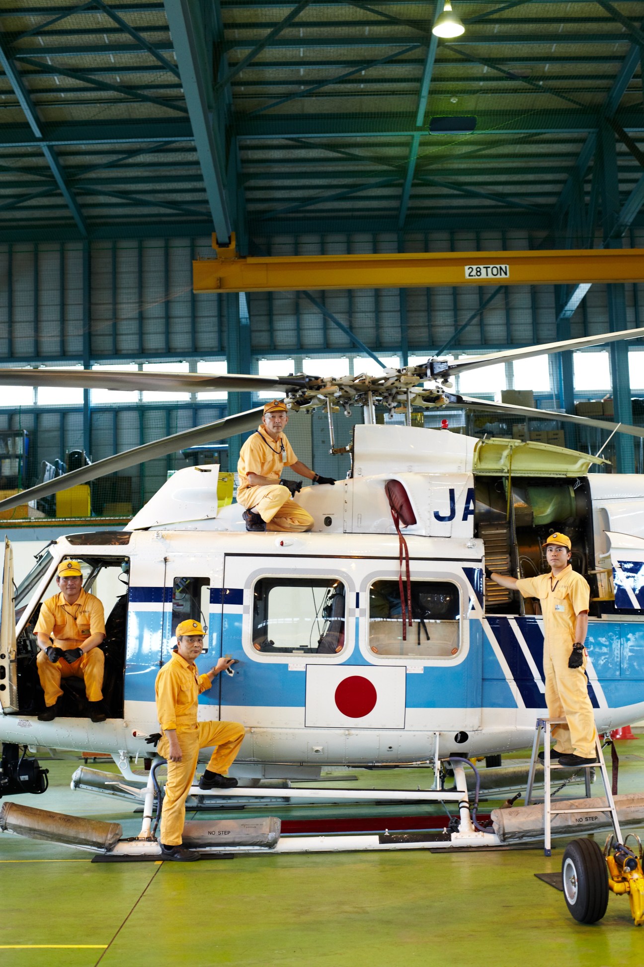 Mechanics in yellow uniforms work on a white and blue Bell 412 helicopter inside a hangar at Hiroshima Air Station.