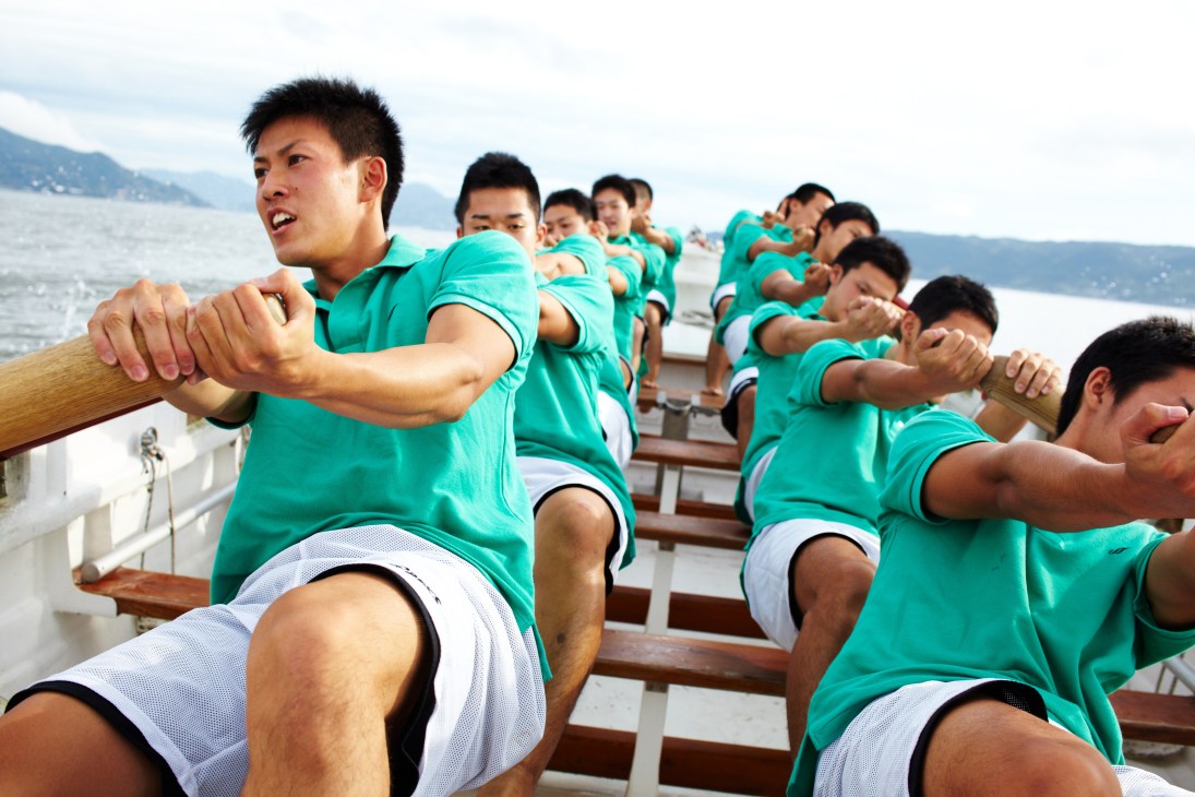 Japan Coast Guard academy students in green shirts rowing together in a wooden cutter boat on the water.