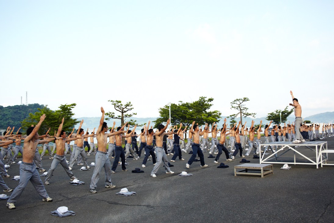 Shirtless Japan Coast Guard academy students perform synchronized morning exercises in formation outdoors.