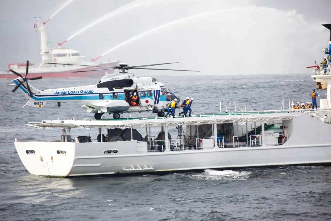 Japan Coast Guard Bell 412 helicopter conducting a hoist-rescue demonstration over a patrol vessel at sea.