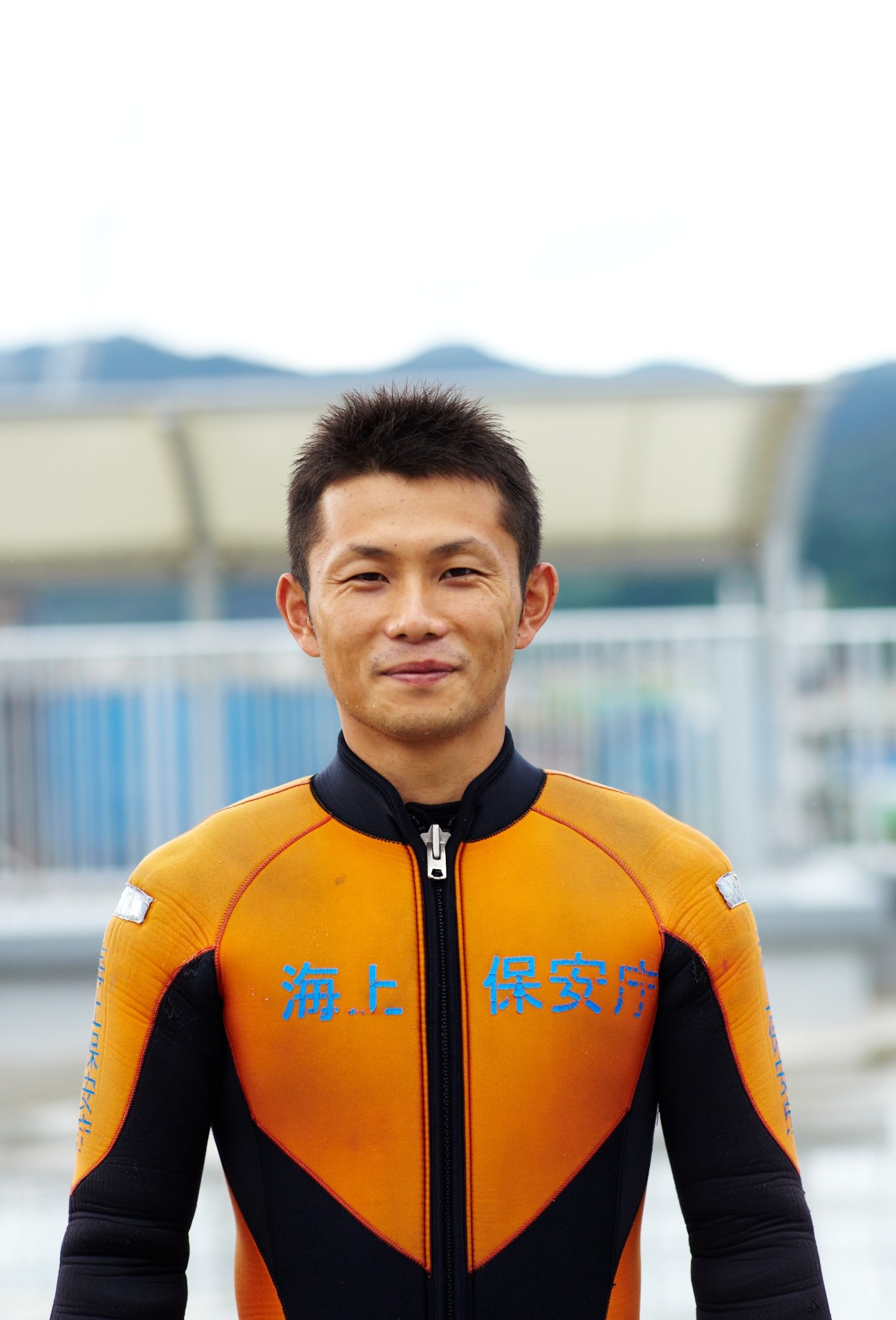 A JCG diving instructor in an orange and black wetsuit stands outdoors near a waterfront facility.