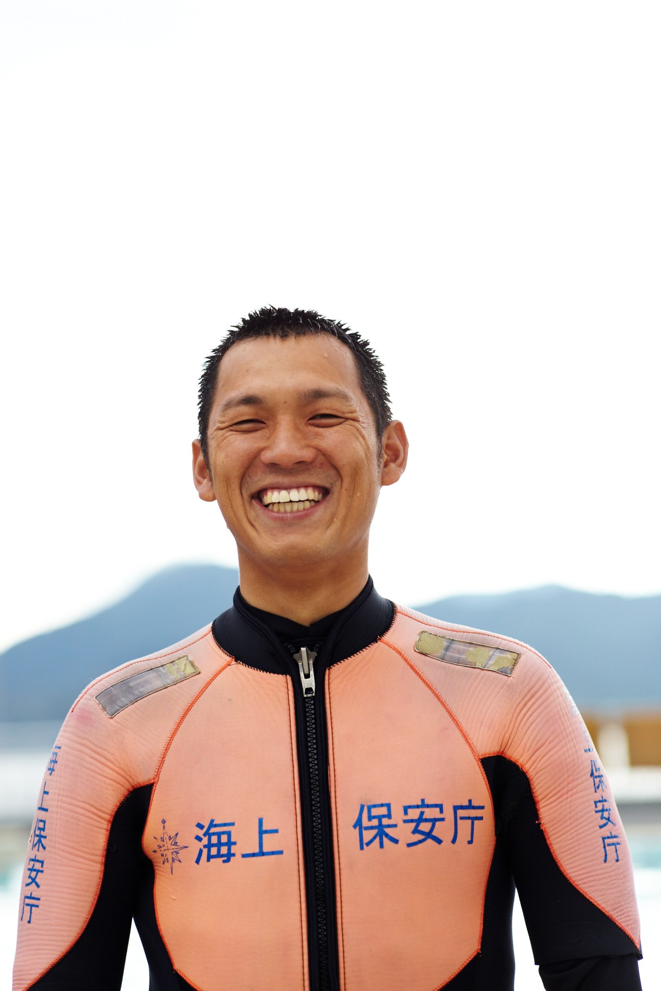 A smiling male Japan Coast Guard diving instructor wearing a coral and black wetsuit with Japanese characters.