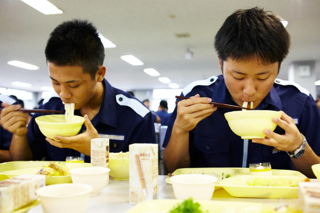 Two Japan Coast Guard academy students in uniforms eating noodles with chopsticks in the canteen.
