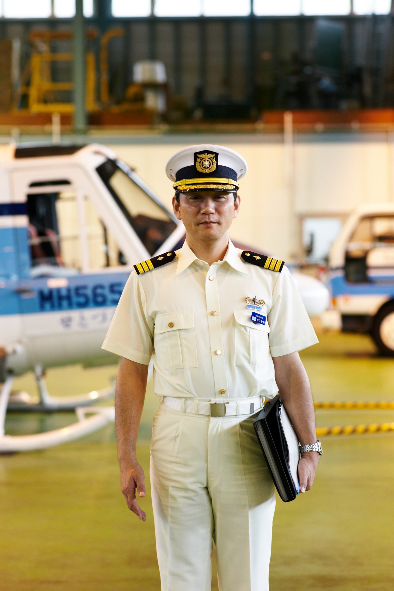 Japan Coast Guard executive officer in white uniform and cap standing in helicopter hangar holding a folder.