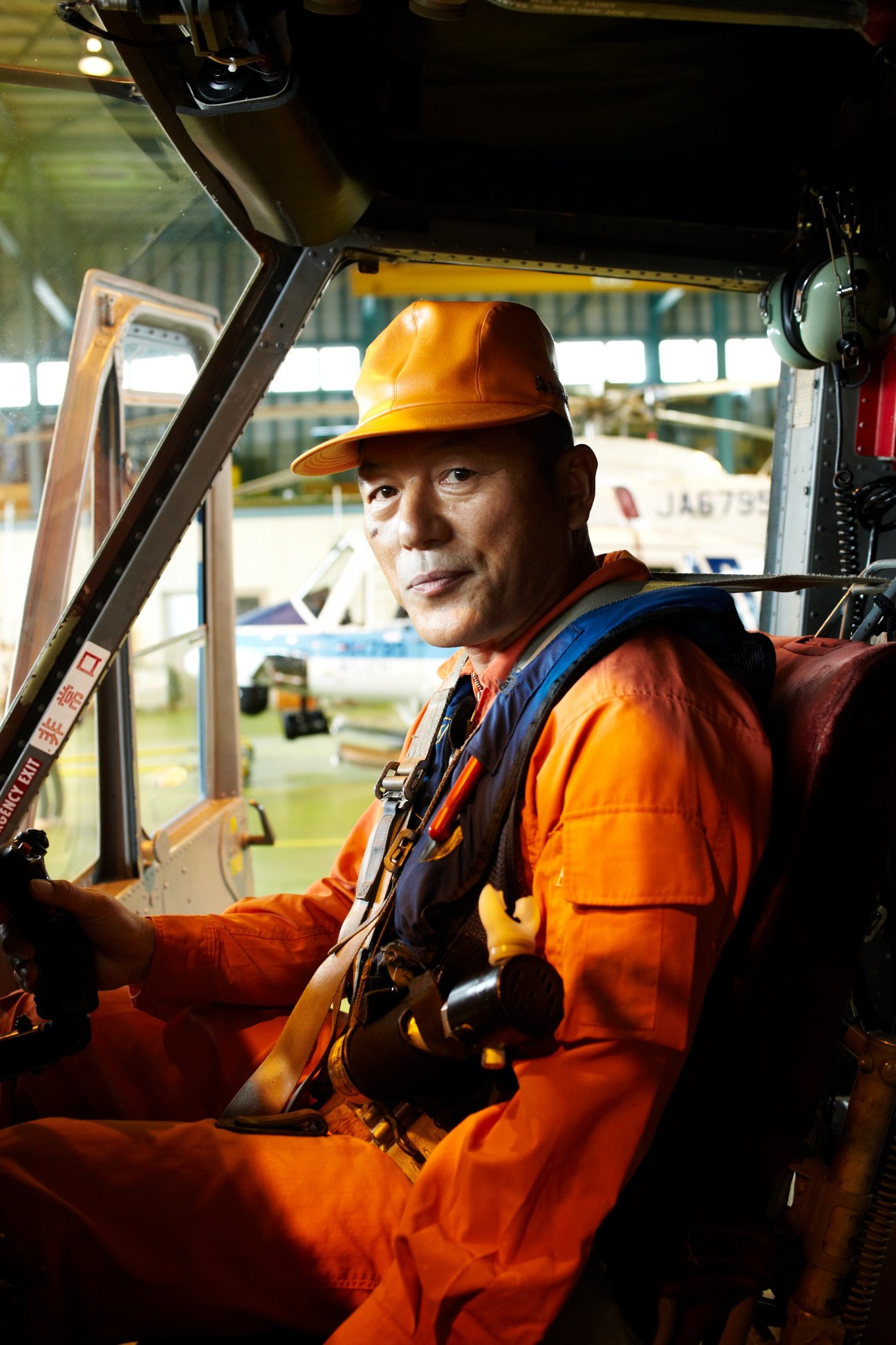 Chief helicopter pilot Haruo Ozaki in orange flight suit and yellow helmet sitting in helicopter cockpit.