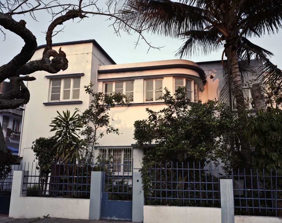 A 1930s Art Deco house in Maputo with cream-coloured facade, curved windows, and blue metal gates, surrounded by tropical trees.