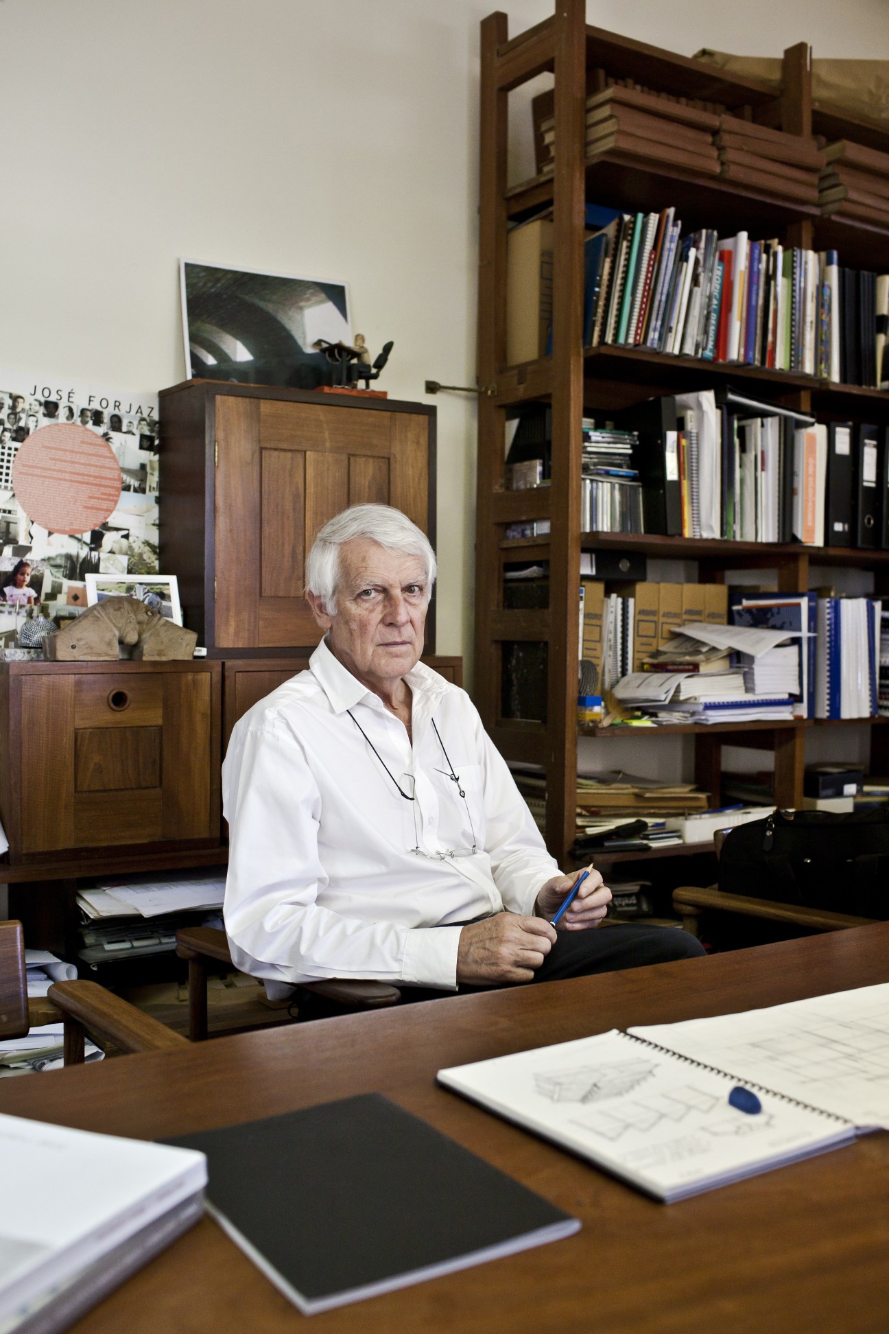 José Forjaz, an elderly man with white hair wearing a white shirt, sits at his wooden desk in his studio surrounded by books, sketches and architectural drawings.
