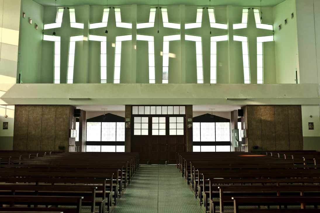 Interior of a modernist church with green walls, geometric skylights, wooden pews, and dark wooden doors.