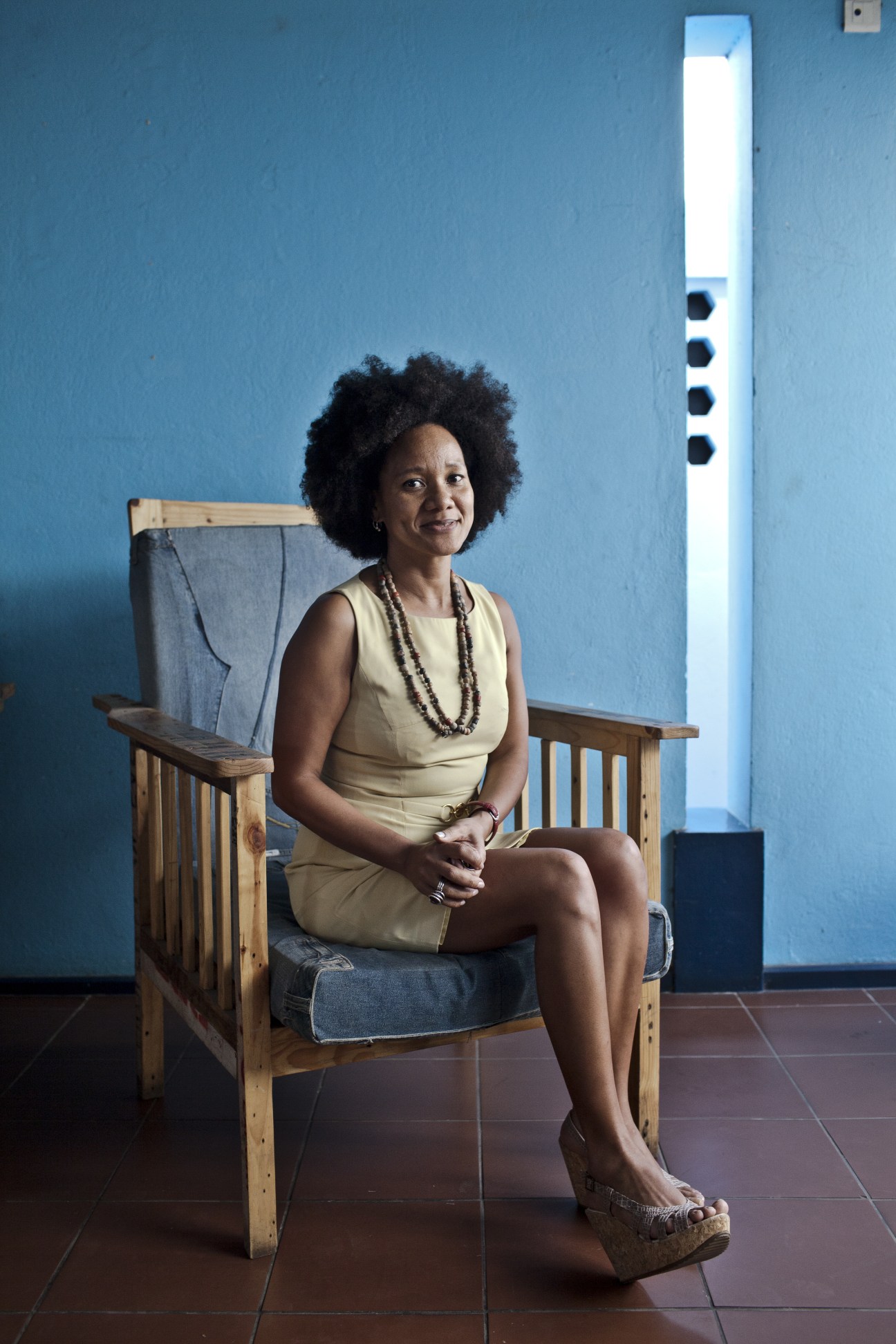 Fashion designer Maria Elisa Chim seated in a wooden chair against a blue wall.