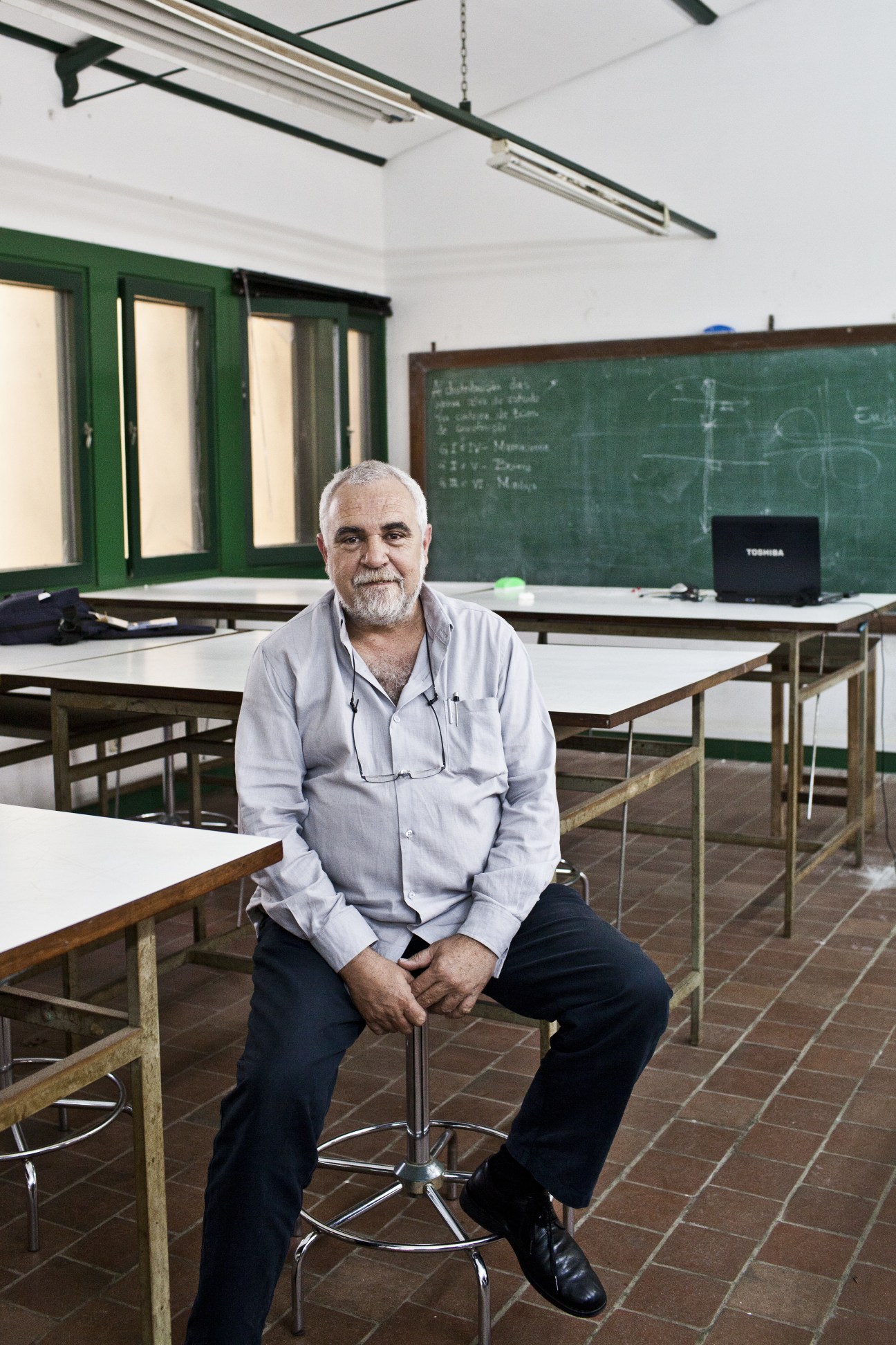 Luis Lage, professor of architecture, sitting in an empty classroom with a chalkboard behind him.