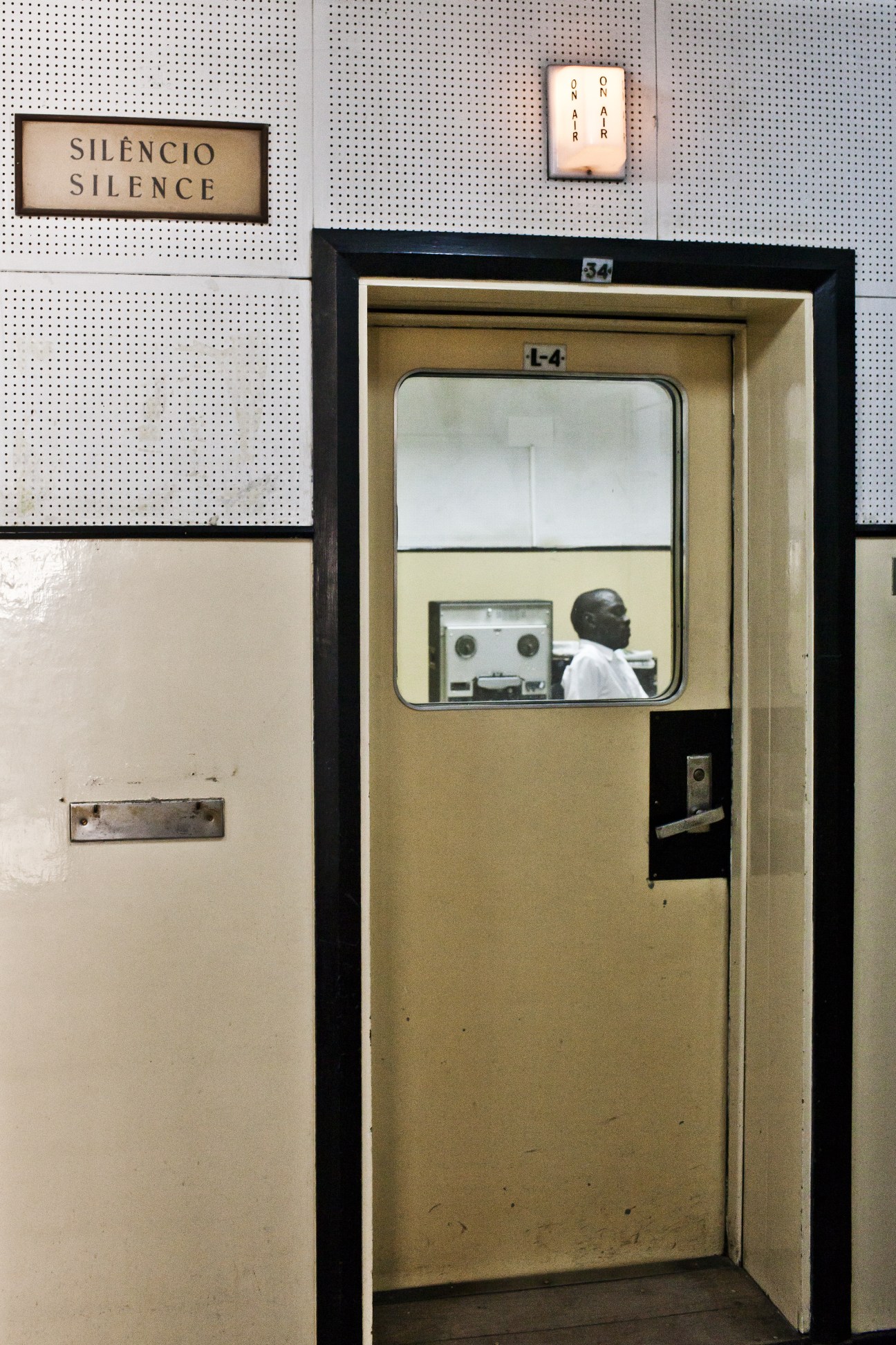 Radio Mozambique studio with man at controls visible through window, silence sign above