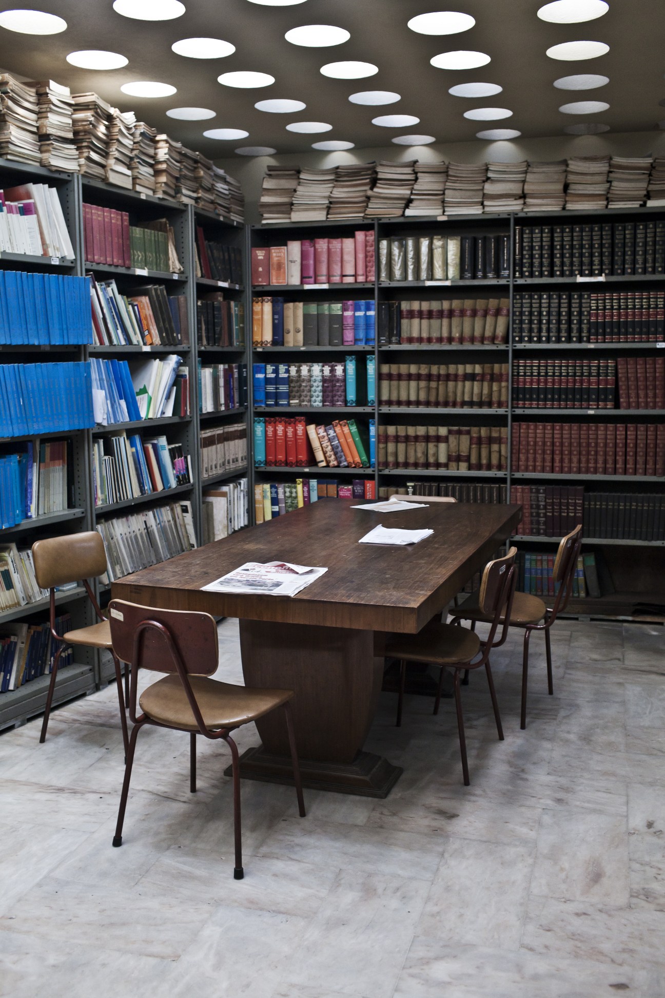 Library interior at Radio Mozambique with floor-to-ceiling bookshelves, wooden table, mid-century chairs and circular ceiling lights.
