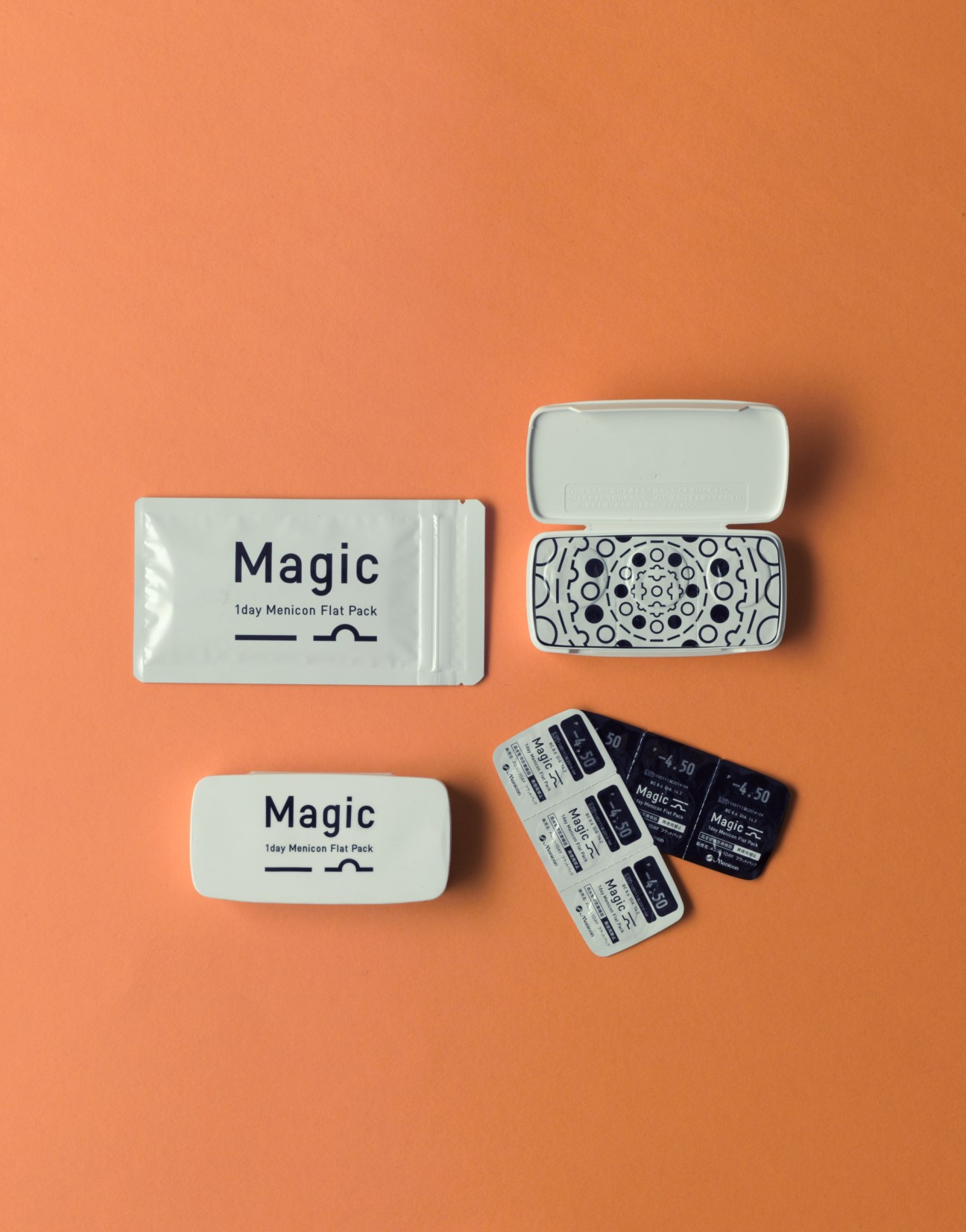Menicon Magic flat contact lens packaging displayed on an orange background, showing opened cases and individual blister packs.