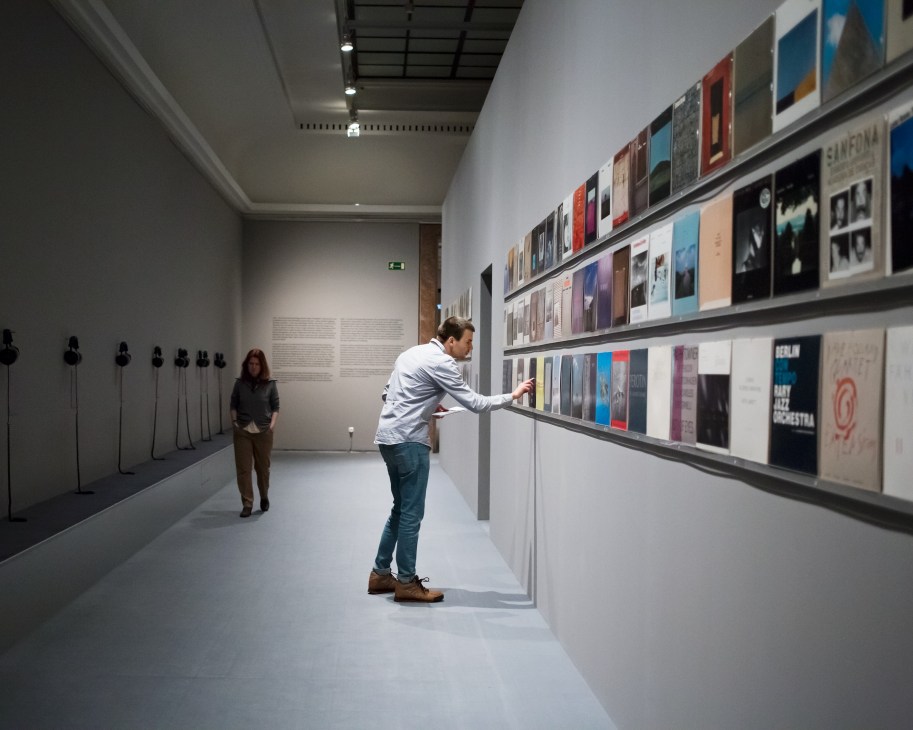 Man examining colorful vinyl record covers displayed on a wall in a gallery space with listening stations.