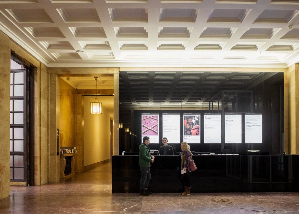 Modernised reception area of Haus der Kunst with gold-leafed walls, coffered ceiling, and black information display panels.