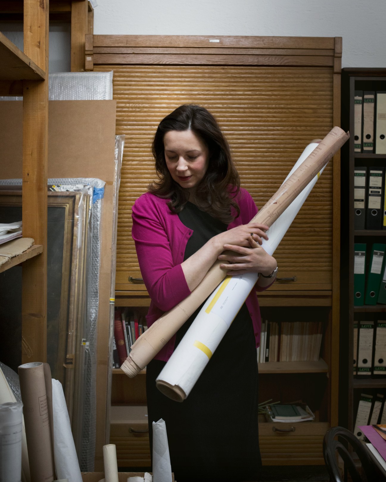 Archivist Sabine Brantl holds a rolled document in Haus der Kunst's archive surrounded by wooden storage units and filed records.