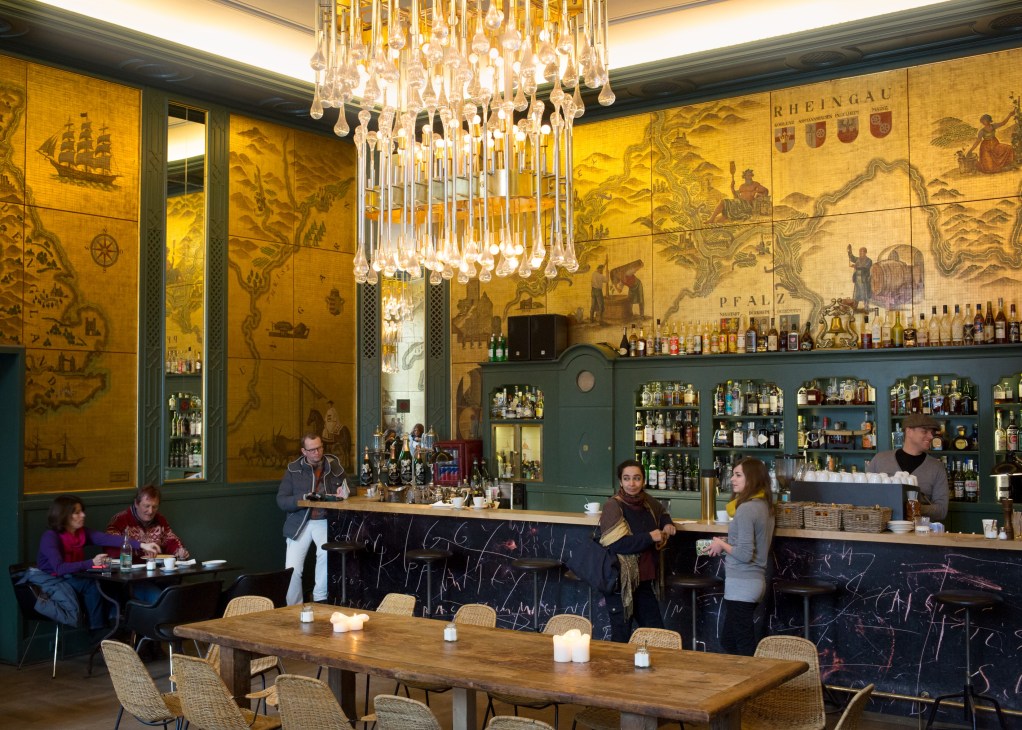 The Golden Bar at Haus der Kunst features yellow wall murals, a crystal chandelier, green cabinetry with bottles, and wooden tables with diners.
