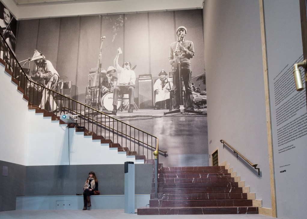 A visitor sits on a bench beside a staircase with a large black and white photograph of musicians performing displayed on the wall above.