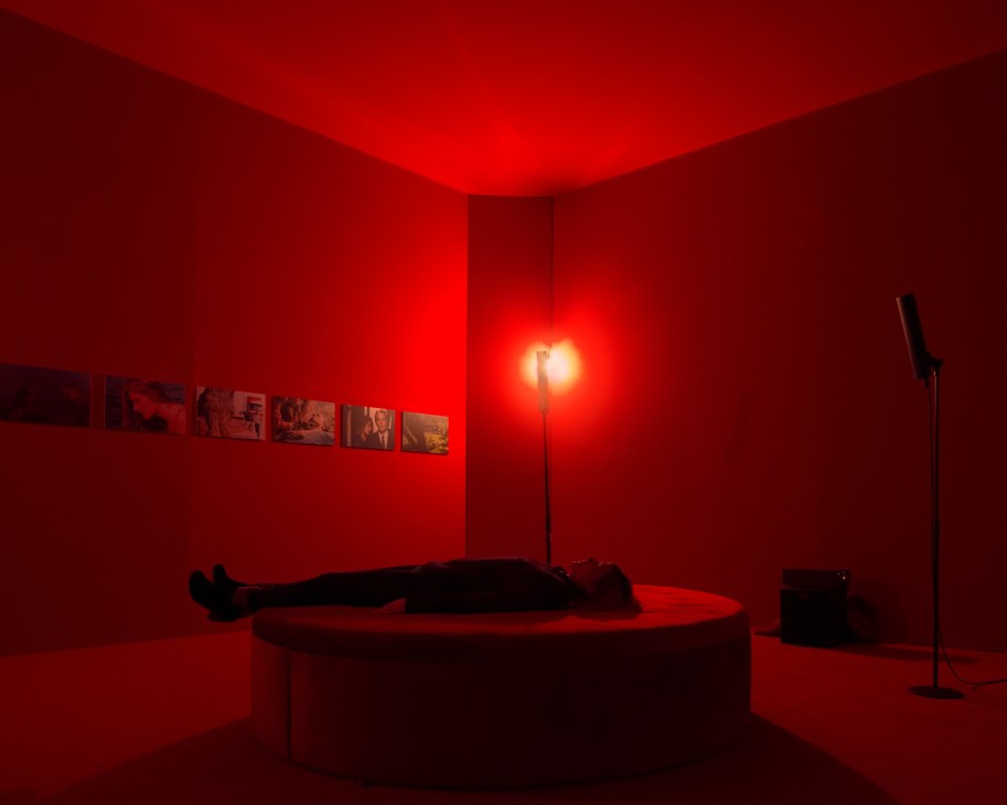 A visitor lies on a platform in a red-lit gallery space listening to music during an ECM record label exhibition.