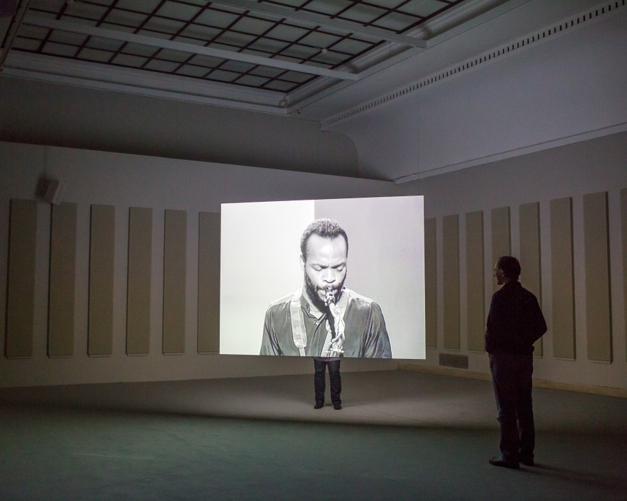 Two visitors watch a black and white video projection of a musician playing saxophone in a gallery space.
