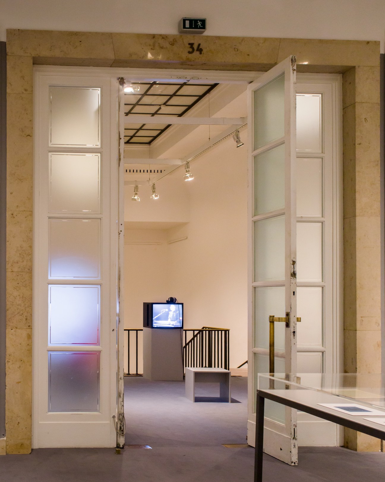 Interior gallery space with white doors, grid ceiling panels, and video monitor displaying the ECM music label exhibition.