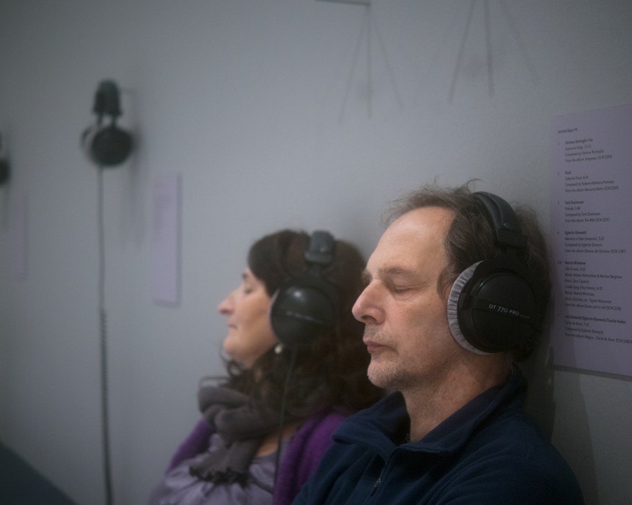 Two visitors wearing headphones listen to music in an exhibition space with text panels on the wall.