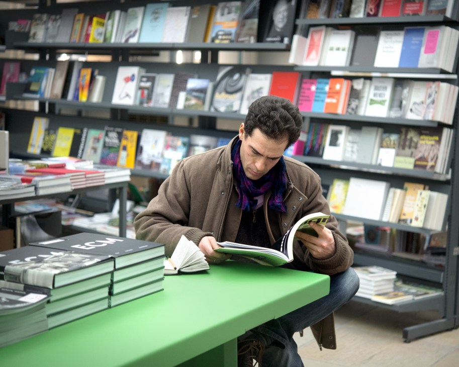 A visitor browses an art book at a green table in Haus der Kunst's bookstore, surrounded by shelves of colorful publications.