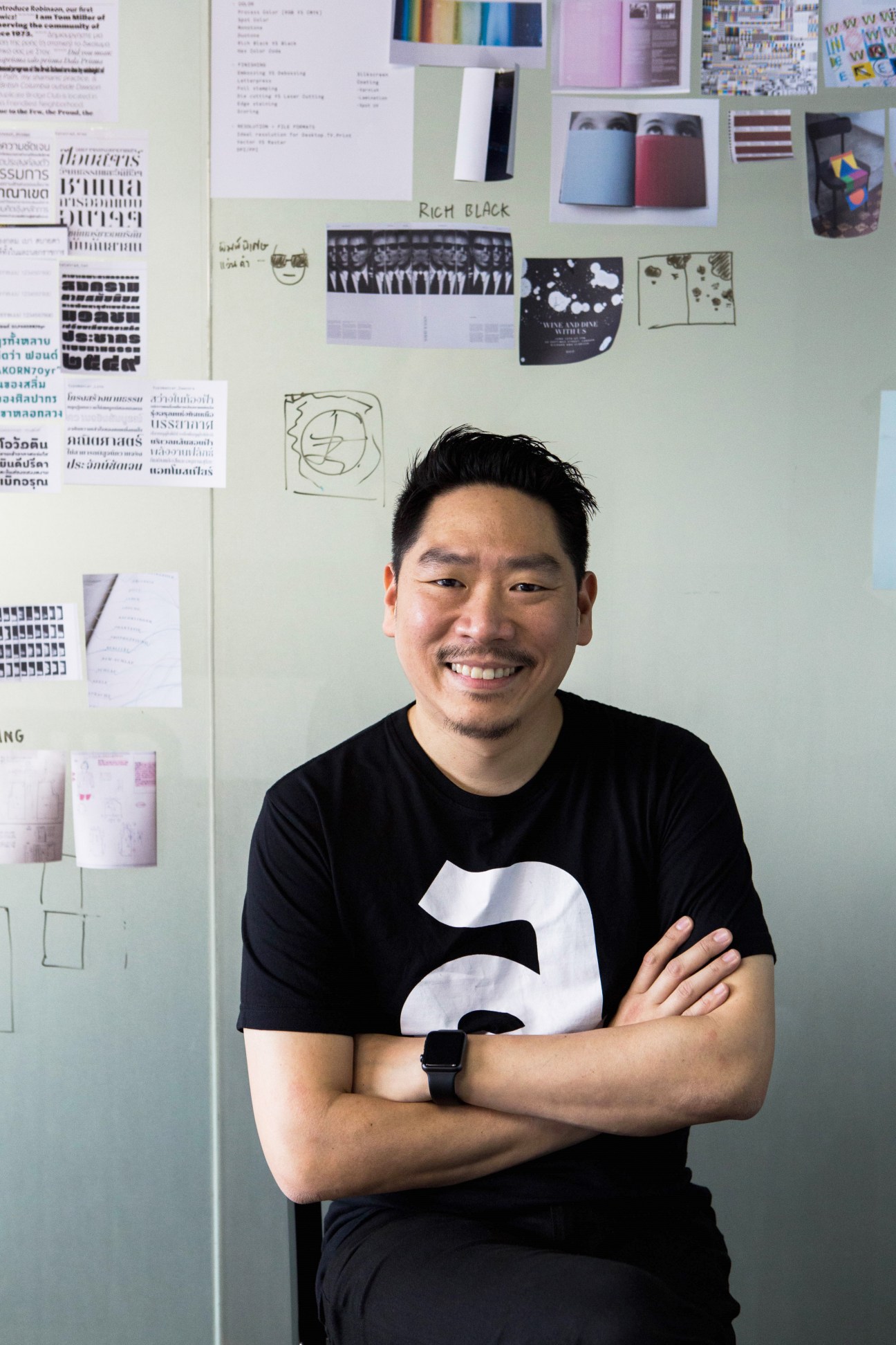 Smiling man with crossed arms in black t-shirt standing against wall covered with design materials and sketches