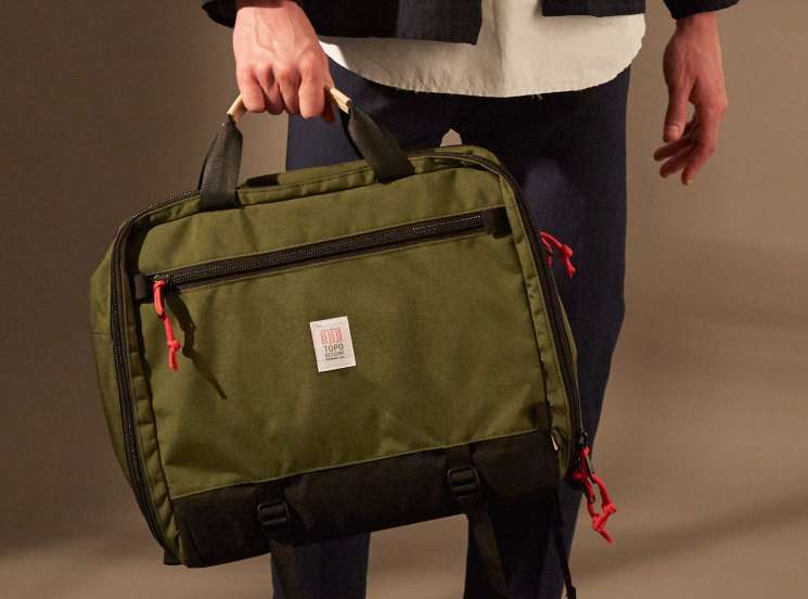Person wearing navy jacket, white shirt, black trousers and white Adidas Stan Smith trainers holding an olive Topo Designs bag.