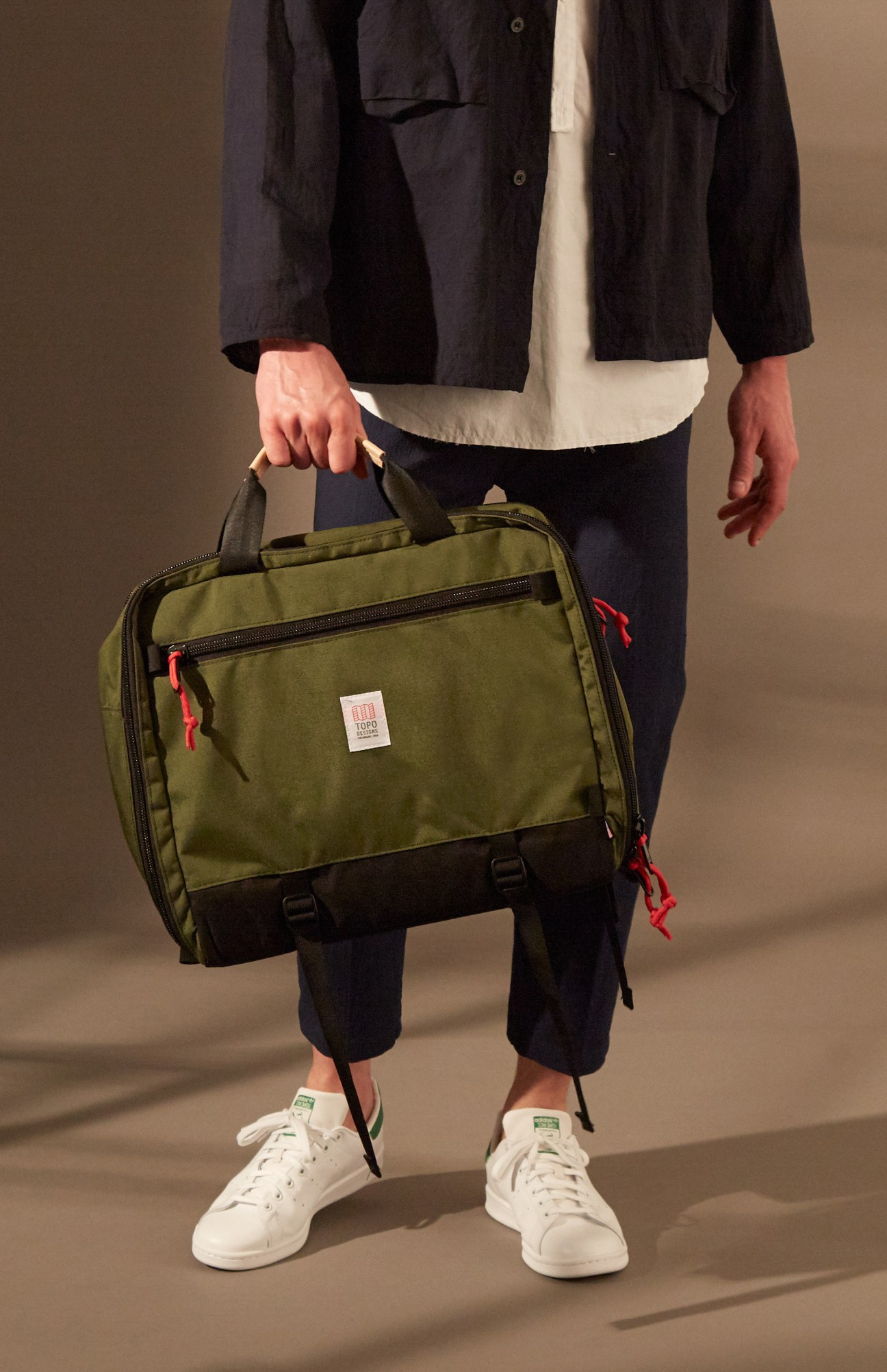 Person wearing navy jacket, white shirt, black trousers and white Adidas Stan Smith trainers holding an olive Topo Designs bag.